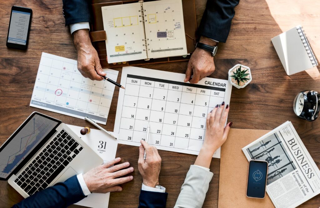 Overhead shot: three people planning with calendars, a laptop, phone, and "BUSINESS" newspaper on a wooden desk.