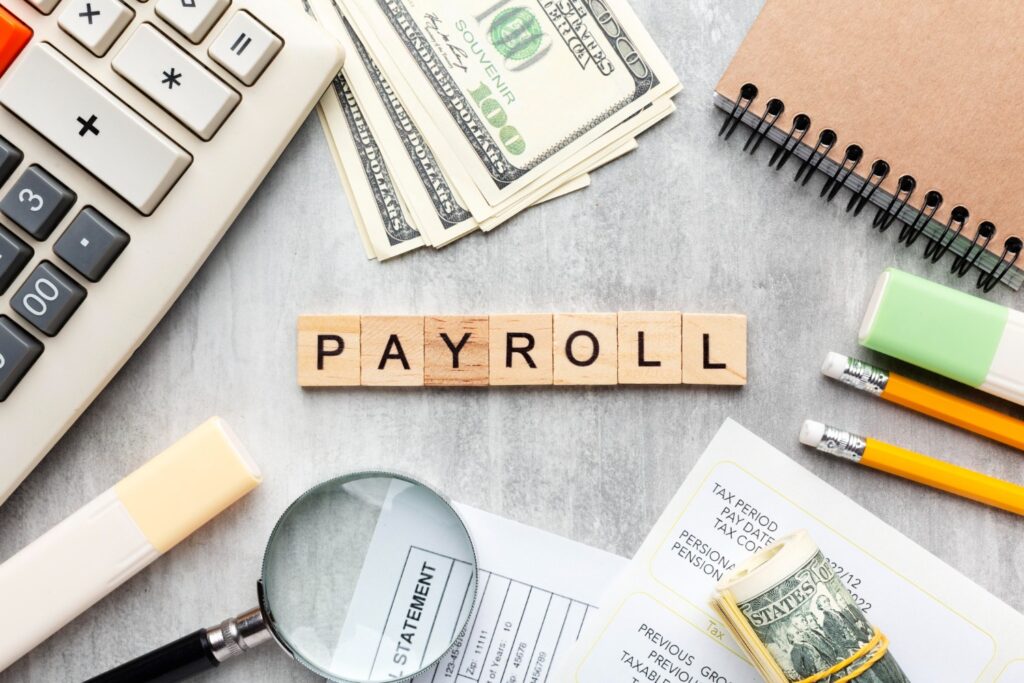 Overhead view of "PAYROLL" spelled out in wooden blocks, surrounded by money, calculator, and financial documents.