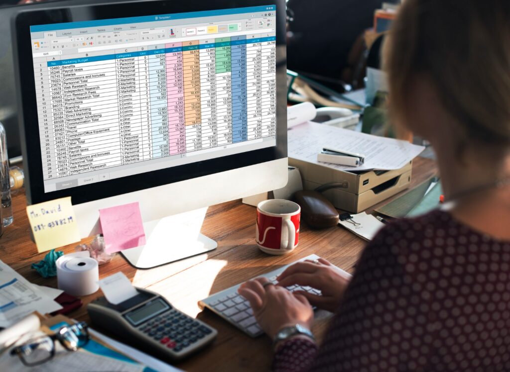 A person works on a computer displaying a detailed financial spreadsheet, with a calculator and coffee mug nearby on a wooden desk.