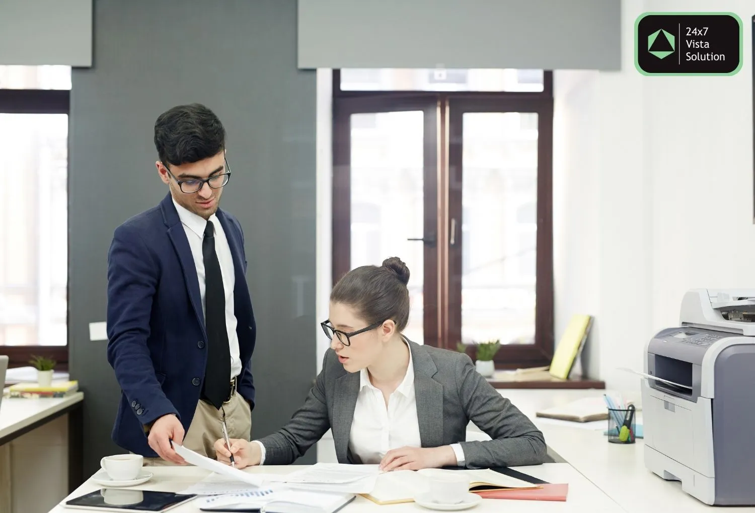 A male and female colleague in business attire are collaborating on a document at a desk in an office.