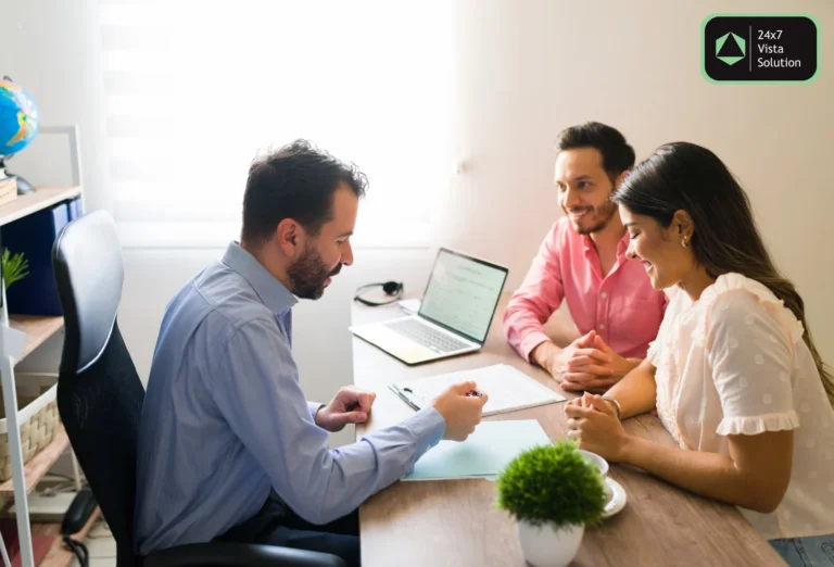 A man is speaking with a smiling couple at a desk, reviewing documents. A laptop, globe, and plant are on the table.