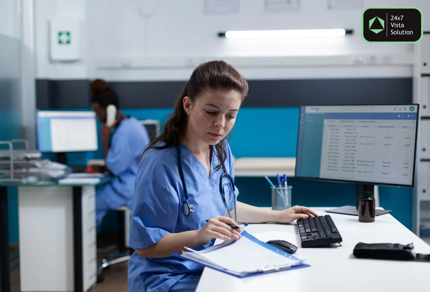 A female nurse in blue scrubs and a stethoscope sits at a desk, looking at a clipboard with papers. She is in an office with a computer monitor displaying data.